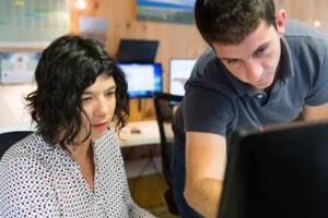 Deux collègues, une femme aux cheveux noirs bouclés et un homme, regardent un écran d'ordinateur ensemble dans un bureau avec des moniteurs allumés en arrière-plan.