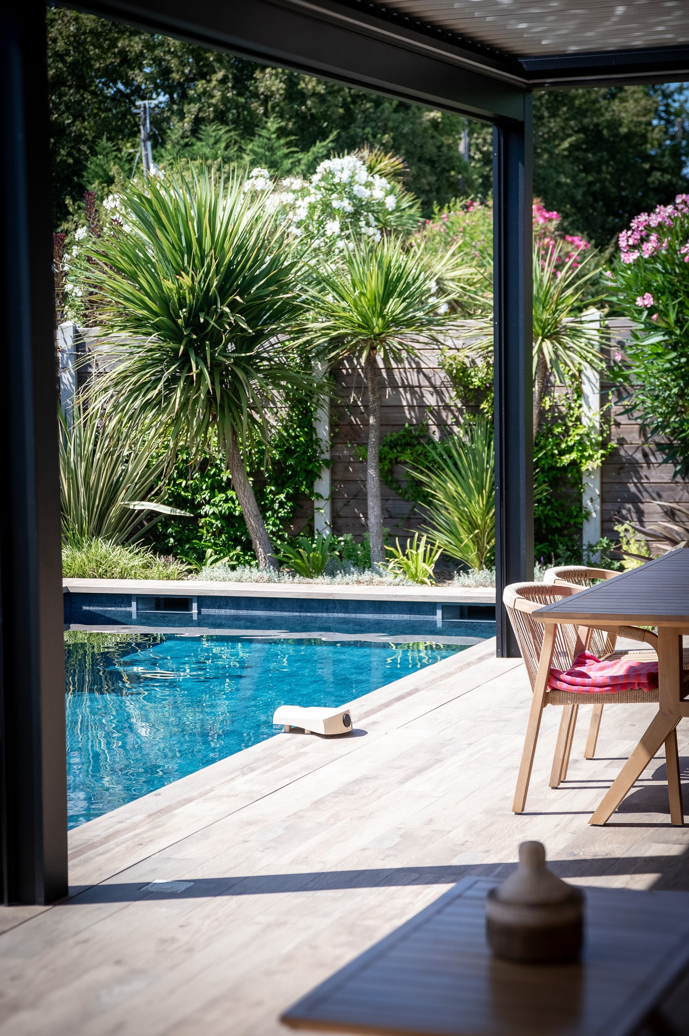 Vue depuis une terrasse couverte sur une piscine turquoise, entourée de palmiers et de végétation luxuriante sous un soleil éclatant.