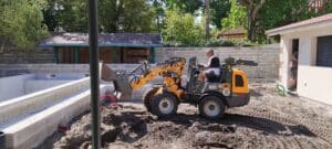 Un ouvrier conduit un chargeur compact Wacker Neuson WL25 sur un chantier de construction de piscine, terre meuble et blocs de béton visibles.