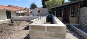 Chantier de construction extérieur avec des murs en parpaings formant une structure rectangulaire (possible bassin) au premier plan, terre battue, et une annexe en bois.