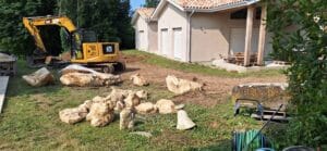 Mini-pelle CAT jaune déplaçant de gros rochers à côté d'une maison neuve avec toit en tuiles rouges et volets roulants